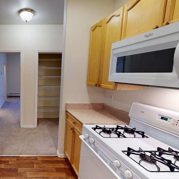 kitchen interior here at the highlands luxury apartments with light wood cabinets, a gas stove, white microwave, and double sink. hallway with open pantry on the right leads to another room.