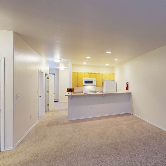 spacious, empty living room here at the highlands luxury apartments with beige carpet and walls, leading to a kitchen with yellow cabinets, white appliances, and warm ceiling lights.