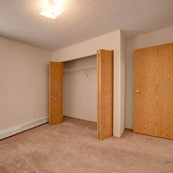 empty bedroom here at aspen terrace apartment homes with beige walls and carpet. a window with blinds on the left, open closet with wooden doors in the center, and a closed door on the right.