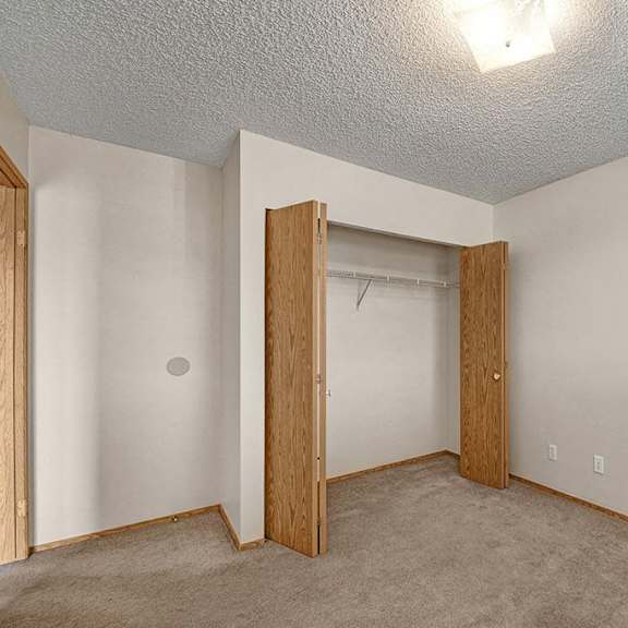empty beige bedroom here at aspen terrace apartment homes with open wooden closet doors revealing a shelf and rod. the room has a carpeted floor, textured ceiling, and a bright light fixture.