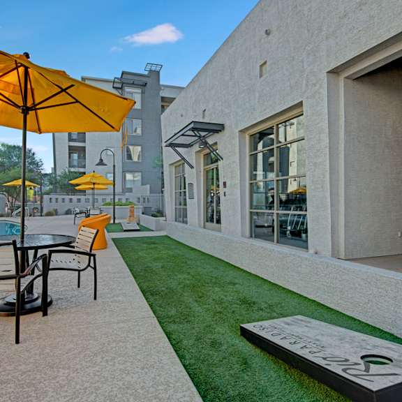 a lively outdoor courtyard here at rio paradiso featuring a poolside patio with round tables and chairs shaded by bright yellow umbrellas, manicured turf with a cornhole game, and a nearby swimming pool, all set against modern apartment buildings with large windows and clean architectural lines designed for casual social gatherings.