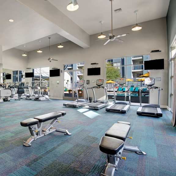 a spacious fitness center here at rio paradiso featuring a wide open layout with treadmills, stationary bikes, weight machines, and adjustable benches arranged on patterned flooring, complemented by high ceilings with ceiling fans, wall-mounted televisions, and large windows that bring in natural light and views of the outdoor pool area.