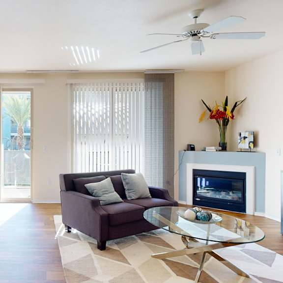 a welcoming open living area here at rio paradiso featuring wood-style flooring, a cozy sofa with accent pillows, a glass coffee table, and a corner fireplace, complemented by a dining nook with a bench and table, ceiling fan overhead, sliding glass doors with vertical blinds, and abundant natural light flowing in from multiple windows.