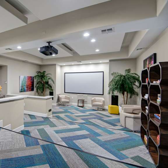 a modern resident lounge here at rio paradiso featuring patterned carpet flooring, a built-in projection screen with ceiling-mounted projector, comfortable lounge chairs, a reception-style counter, decorative wall art, potted greenery, and a shelving area with books and games, all illuminated by recessed lighting for a relaxed communal atmosphere.