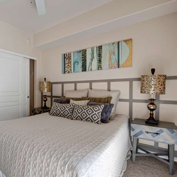 a serene bedroom here at rio paradiso featuring a spacious bed dressed in neutral bedding with patterned accent pillows, matching bedside tables with decorative lamps, and soft carpet flooring. the room includes a ceiling fan, an open closet with shelving, and a private doorway leading to an adjoining bathroom for added comfort and convenience.