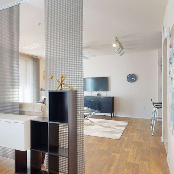 a modern open bedroom here at rio paradiso featuring a stylish shelving divider with textured panel screens that separate the sleeping area from the living space, complemented by wood-style flooring, soft natural light from large windows with vertical blinds, contemporary décor accents, and a clear view into the living room with seating, media console, and wall art.