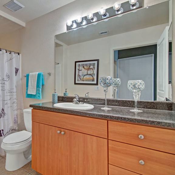 a clean bathroom here at rio paradiso featuring a long wood-tone vanity with a single sink, dark countertop, and wide mirror with overhead lighting, paired with a tub and shower combination dressed in a patterned curtain, neutral tile flooring, a toilet, neatly hung towels, and soft wall art that adds a welcoming touch.