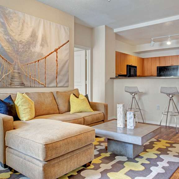 a comfortable living room here at rio paradiso featuring a plush sectional sofa with yellow and navy accent pillows, a modern square coffee table with decorative vases, and patterned area rug over wood-style flooring. the space opens to a kitchen breakfast bar with seating, while large wall art and soft neutral tones create a relaxed, welcoming feel.