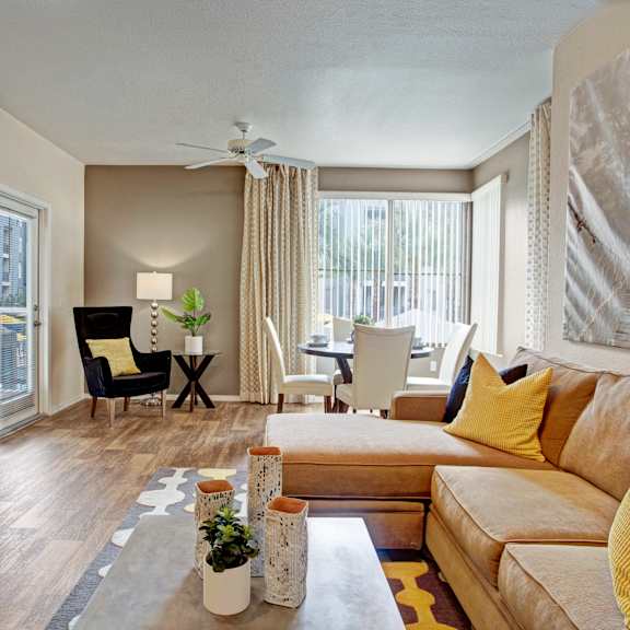 a modern open living room here at rio paradiso featuring wood-style flooring, a plush sectional sofa with accent pillows, a rectangular coffee table with decorative accents, and a wall-mounted television above a low media console, flowing into a dining area with a round table, upholstered chairs, large windows with vertical blinds, and a ceiling fan providing natural light and comfort.