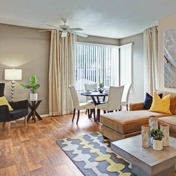 a warm, inviting living room here at rio paradiso featuring a plush sectional sofa with yellow accent pillows, a modern coffee table with decorative accents, and wood-style flooring. the space flows into a cozy dining area near large windows with patterned curtains, while wall art, soft lighting, and neutral décor create a comfortable, welcoming atmosphere.