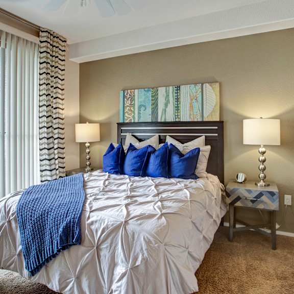 a welcoming bedroom here at rio paradiso featuring a neatly made bed with crisp white bedding and blue accent pillows, a dark wood headboard framed by matching bedside tables with modern lamps, soft carpet flooring, and a sliding glass door with vertical blinds and patterned curtains that bring in natural light from a private balcony.