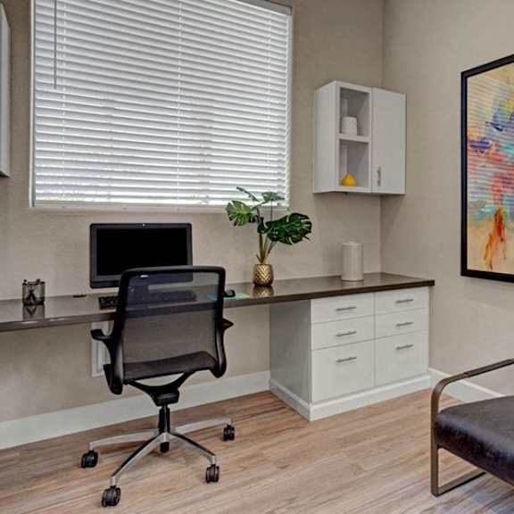 a quiet business center here at sonoma landing featuring a built-in desk with a computer workstation, rolling office chair, and light wood-style flooring, complemented by white cabinetry for storage, a framed abstract art piece on the wall, a comfortable accent chair, and large window blinds that bring in soft natural light.