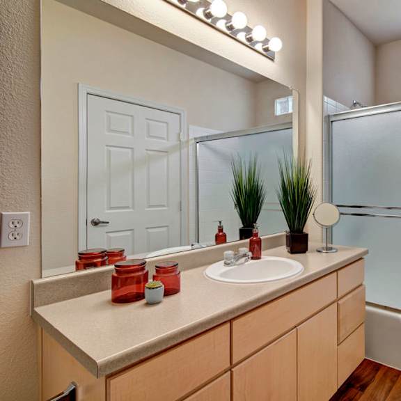 a bright bathroom here at sonoma landing featuring a long vanity with light wood cabinetry, a single sink set into a neutral countertop, and a wide wall mirror with overhead lighting, complemented by decorative greenery, a toilet, and a tub and shower combination enclosed by sliding glass doors, with warm-toned flooring completing the space.