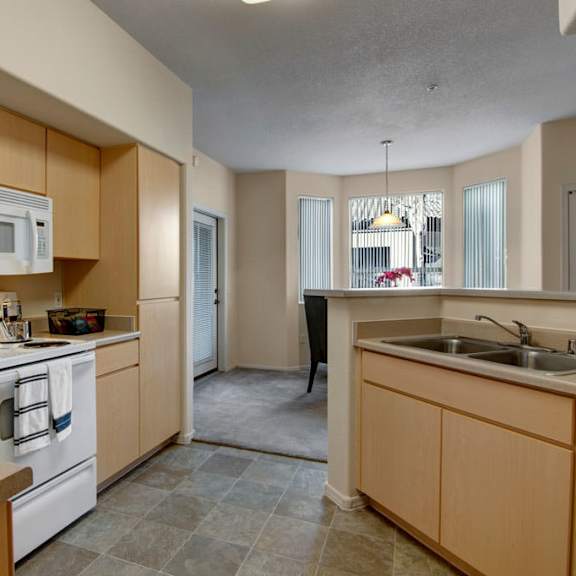 a practical kitchen here at sonoma landing featuring light wood cabinetry, a full appliance setup including a refrigerator, range, microwave, and dishwasher, and wraparound countertops with a double-basin sink, complemented by wood-style flooring, neutral walls, and an open pass-through that connects the kitchen to the adjacent living space.
