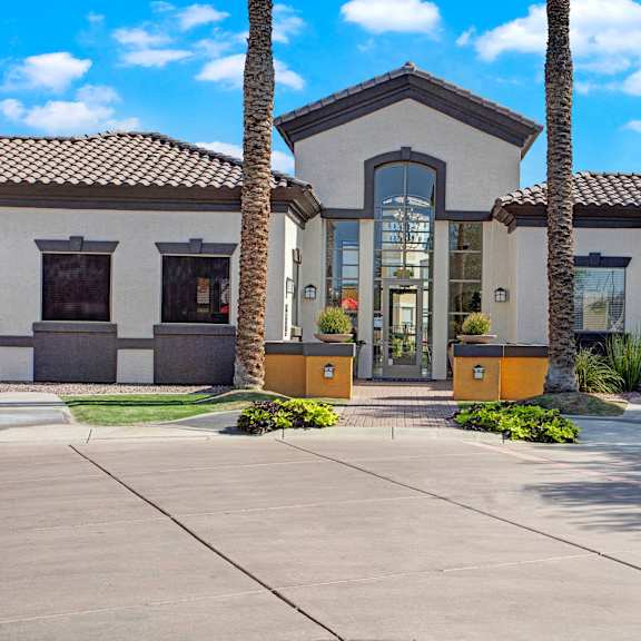 a welcoming leasing office exterior here at sonoma landing featuring a modern stucco building with a tiled roof, tall glass entry doors, and symmetrical architectural lines, framed by mature palm trees, manicured landscaping, decorative planters, and a paved walkway that leads to the main entrance under a bright arizona sky.