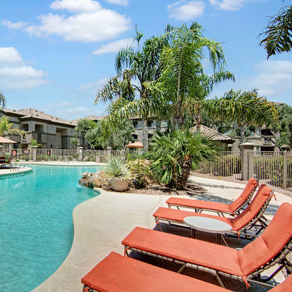 a resort-style outdoor swimming pool here at sonoma landing featuring a curved turquoise pool surrounded by red cushioned lounge chairs, palm trees, and desert landscaping, with shaded umbrellas, a fenced deck, and warm stucco apartment buildings creating a relaxing courtyard setting under a bright blue sky.