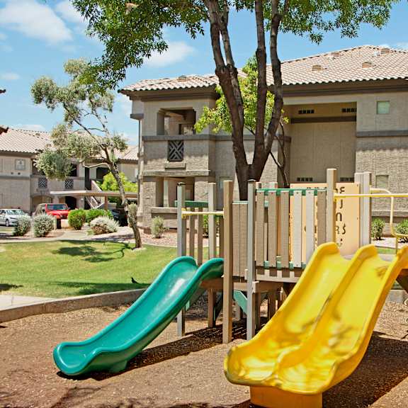 a shaded outdoor playground area here at sonoma landing featuring a compact play structure with bright yellow and green slides, a spiral climbing pole, and a raised platform set on a natural ground surface, surrounded by mature trees, grassy areas, and warm-toned apartment buildings that create a family-friendly community setting.