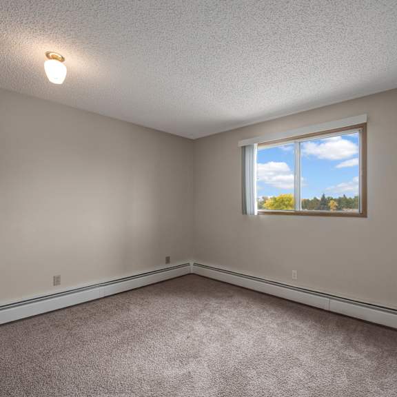 a bright, empty bedroom here at gateway square with soft beige walls, plush carpet flooring, and a large window with vertical blinds letting in natural light and peaceful outdoor views.