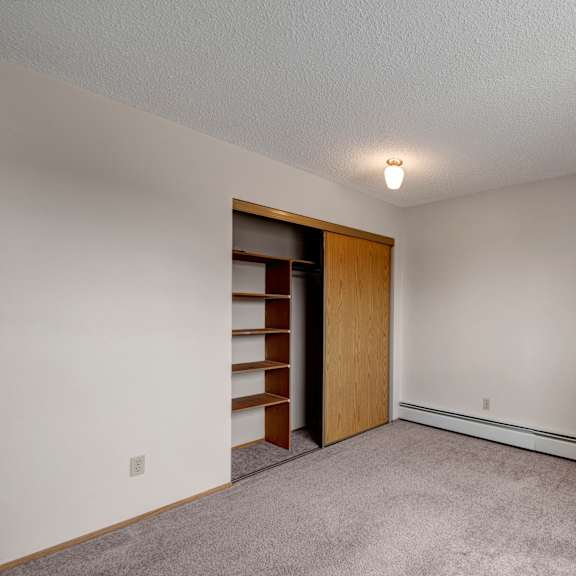 a spacious, empty bedroom here at gateway square with soft beige walls, plush carpet flooring, and a large closet featuring sliding wood doors and built-in shelving for convenient storage.