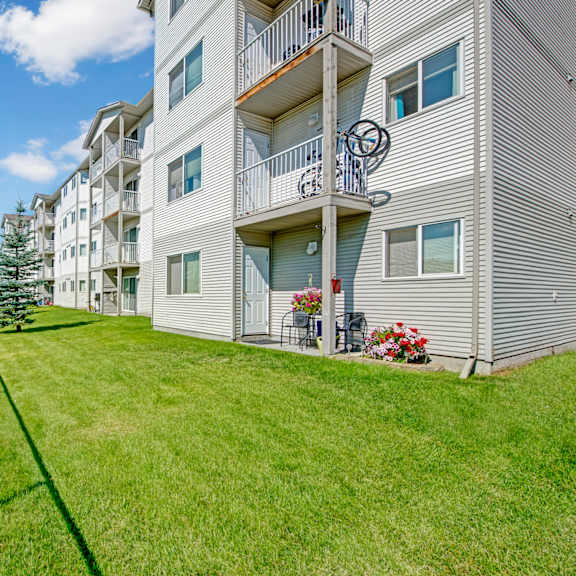 a landscaped exterior view here at sonora showcasing a well-maintained green lawn alongside a multi-story apartment building with light-colored siding, private balconies on each level, patio seating with potted flowers, mature trees, and a fenced perimeter under a bright blue sky.