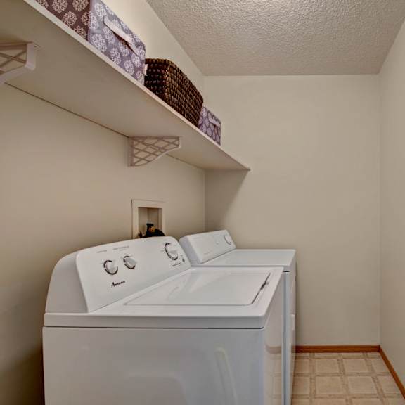 a convenient in-home laundry area here at sonora featuring a full-size washer and dryer, built-in open shelving for storage baskets and supplies, neutral walls, and durable flooring that provides a practical and organized space for everyday laundry needs.