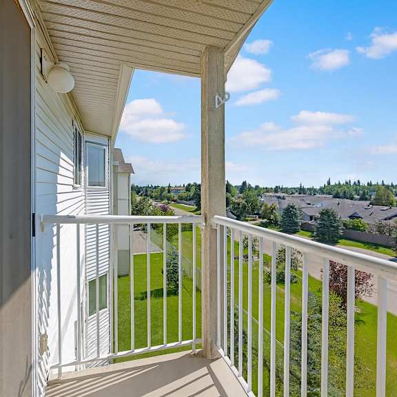 a private outdoor balcony here at sonora featuring white railing, durable concrete flooring, and an elevated view overlooking lush green landscaping, mature trees, and a peaceful residential neighborhood under bright blue skies, offering a relaxing space to enjoy fresh air and scenic surroundings.
