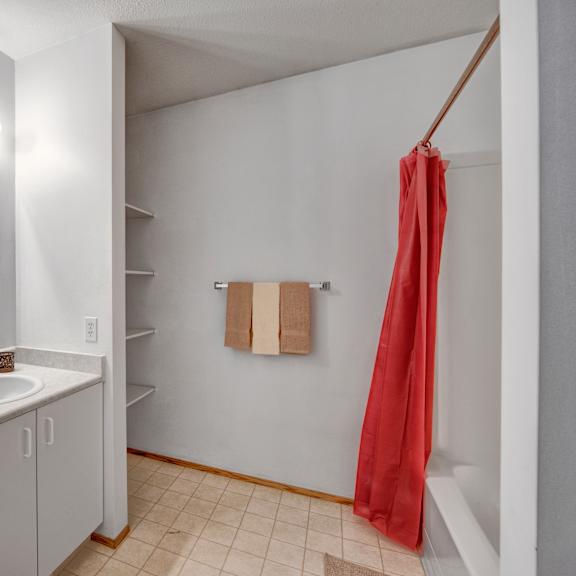 a clean bathroom vanity area here at willowview terrace with a white sink countertop, a large wall mirror, and bright overhead lighting. open shelving provides extra storage space, while a towel bar and a simple tub-and-shower combo with a red shower curtain complete the practical layout.