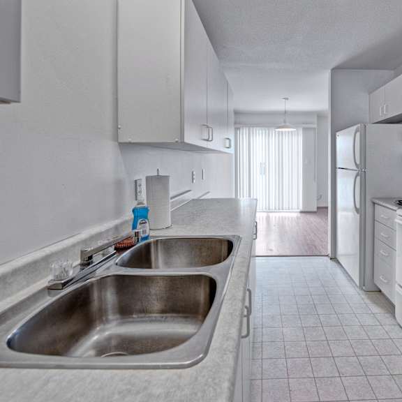 a bright galley-style kitchen here at willowview terrace with white cabinets, light countertops, and a double stainless steel sink. white appliances including a stove, range hood, and refrigerator line the space, with durable tile flooring that leads into the dining area with a sliding glass door for natural light.