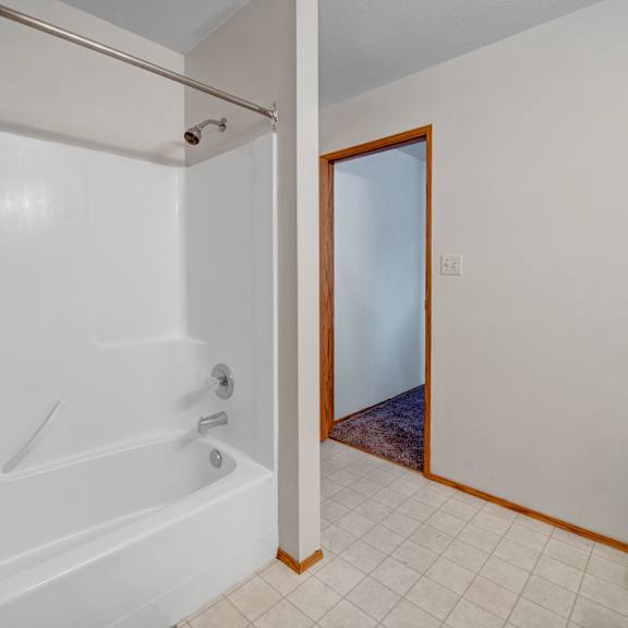 a bright bathroom tub and shower area here at willowview terrace with smooth white surround walls, a simple showerhead, and a sturdy curtain rod. light tile flooring and warm wood trim lead out to the hallway, keeping the space clean, open, and easy to maintain.
