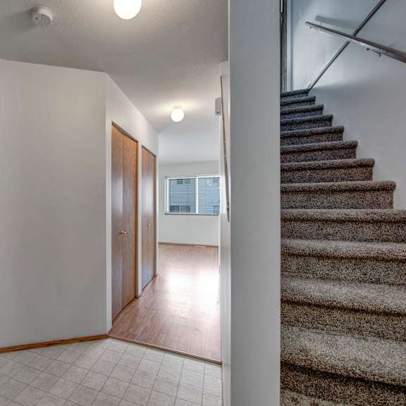 a bright entry hallway here at willowview terrace with light tile flooring, warm wood closet doors, and a clear walkway leading into the main living area. carpeted stairs with a simple handrail add easy access to the upper level, while the nearby kitchen cabinetry keeps the layout practical and open.