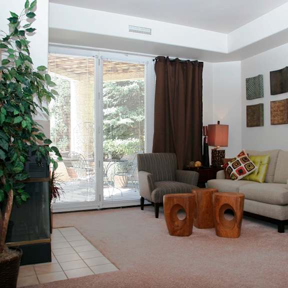 a cozy inviting living room here at cheyenne creek luxury featuring a comfortable sofa with accent pillows, a patterned armchair, and sculptural wooden side tables arranged on plush carpet, with a fireplace, abundant greenery, and large sliding glass doors that open to a private patio, creating a warm, relaxed, and welcoming atmosphere filled with natural light.