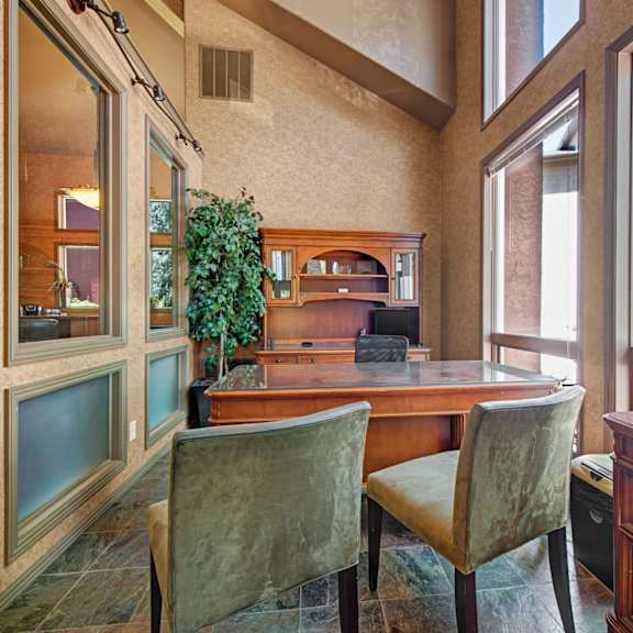 a bright business center here at cheyenne creek luxury featuring a polished wooden desk with upholstered guest chairs, a built-in hutch workspace, large floor-to-ceiling windows, textured walls, and natural light that creates a quiet, professional environment for working or studying.