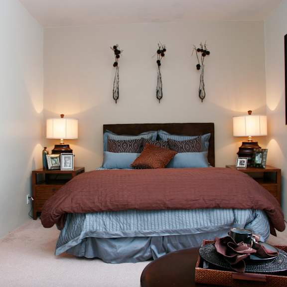 a serene bedroom here at cheyenne creek luxury featuring a centered bed with layered blue and brown bedding, matching wooden nightstands with table lamps, soft carpet flooring, and neutral walls accented by decorative wall art, with an open view into an adjoining bathroom and a small accent table in the foreground.