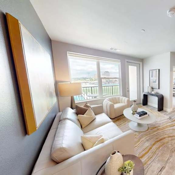 a contemporary living room here at experience at epicenter showcasing wood-style flooring, a light neutral sofa with accent pillows, a matching armchair, a round coffee table styled with decor, an abstract area rug, a floor lamp, framed wall art, open shelving with decorative accents, and large windows with a glass door that fill the space with natural light and offer balcony views.