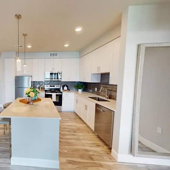 a contemporary kitchen here at experience at epicenter showcasing gray shaker-style cabinetry, stainless-steel appliances, a white quartz island with a decorative centerpiece, pendant lighting, a hexagon tile backsplash, recessed ceiling lights, and warm wood-style flooring that creates a clean, modern, and inviting cooking space.