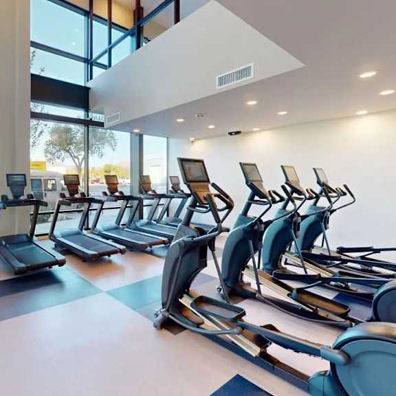 a modern fitness center here at experience at epicenter featuring rows of cardio equipment, including treadmills and elliptical machines, arranged on sleek flooring beneath high ceilings. floor-to-ceiling windows fill the space with natural light and showcase mountain views, creating an open, energizing environment for workouts.