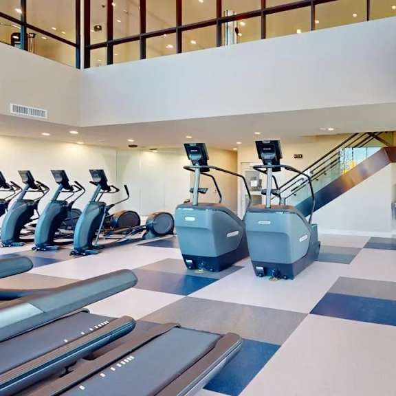 a spacious fitness center here at experience at epicenter featuring a wide selection of cardio equipment, including treadmills, ellipticals, and stair machines, along with strength and free-weight stations. high ceilings, an open mezzanine, modern lighting, and expansive windows create a bright, motivating environment designed for a full-body workout experience.
