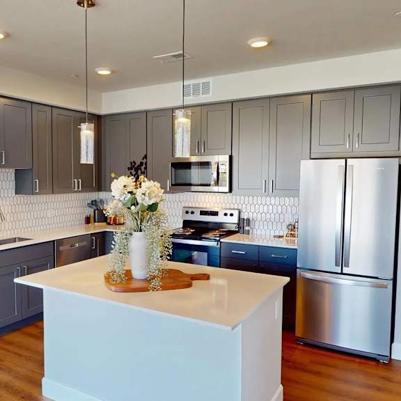 a contemporary kitchen here at experience at epicenter featuring gray shaker-style cabinets, white countertops, and a large center island with a smooth surface, accented by pendant lighting overhead. stainless-steel appliances, including a refrigerator, stove, microwave, dishwasher, and sink, pair with a patterned tile backsplash and warm wood-style flooring.