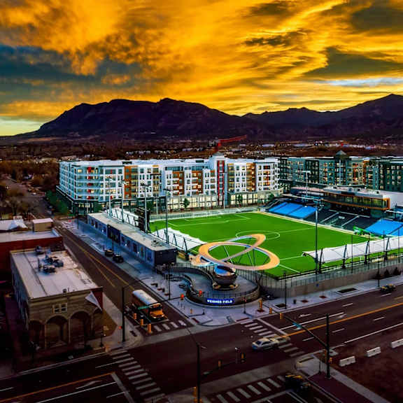 a dramatic aerial view here at experience at epicenter showcasing a vibrant urban neighborhood centered around a professional-style sports field, surrounded by modern apartment buildings and illuminated streets. the scene is framed by mountain backdrops and a glowing sunset sky, highlighting the community’s energetic, city-connected setting.