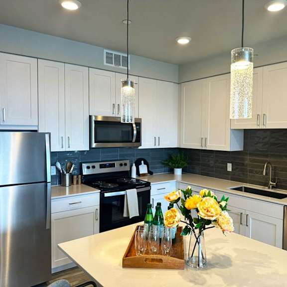 a modern kitchen here at experience at epicenter showcasing white shaker-style cabinets, light countertops, and a spacious center island with decorative accents and pendant lighting above. stainless-steel appliances, including a refrigerator, stove, microwave, dishwasher, and sink, pair with a dark tile backsplash, under-cabinet lighting, and warm wood-style flooring.