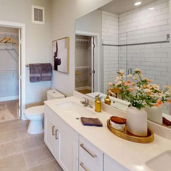 a modern bathroom here at experience at epicenter featuring light tile flooring, a white vanity with dual sinks, and a wide mirror reflecting bright recessed lighting. a walk-in shower with white subway tile sits beside a clean-lined toilet, while an open walk-in closet with shelving and hanging space adds convenient storage.