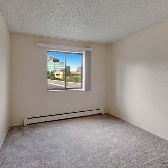 empty beige bedroom here at the continental apartments with textured walls and carpeted floor. a window reveals a bright, sunny view of modern buildings and greenery outside.