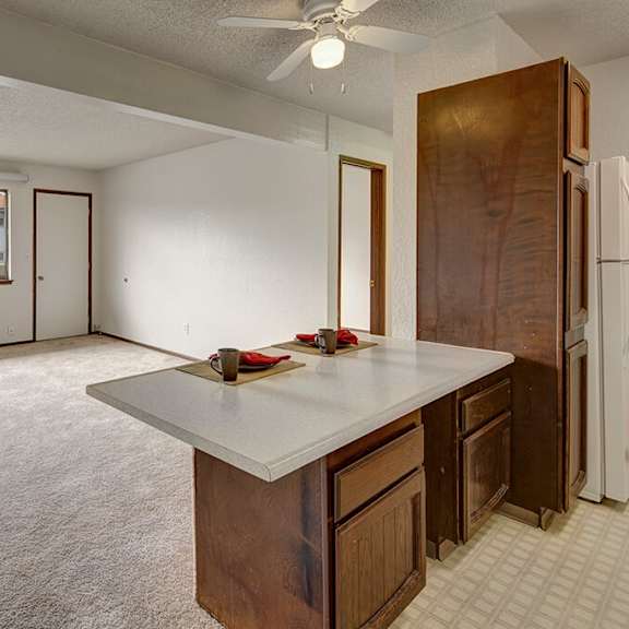 spacious, empty living room here at the continental apartments with beige carpet and white walls. large window with vertical blinds, overlooking adjacent buildings. open kitchen counter with two place settings.