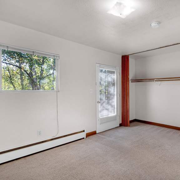 living room here at country lane apartments with beige carpet and white walls. a window with trees outside, a closet with a wooden rod, and a door with blinds are visible, creating a calm, spacious atmosphere.