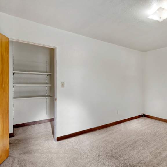 empty bedroom here at country lane apartments with beige carpet, white walls, and a wooden baseboard. an open closet with shelves is visible, and a ceiling light softly illuminates the space.