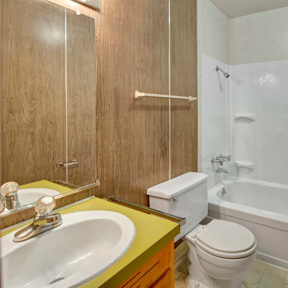 small bathroom here at country lane apartments with a white bathtub and shower, wood-paneled walls, and a yellow countertop with a sink. the atmosphere is simple and clean.