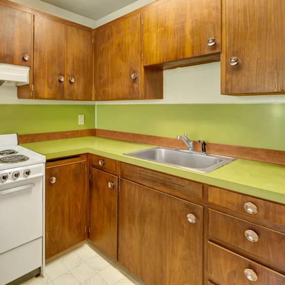 retro kitchen here at country lanes apartments with brown wooden cabinets and a green countertop. white electric stove and stainless steel sink create a vintage feel. cozy and nostalgic ambiance.