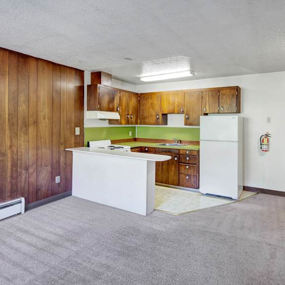 retro-style kitchen here at country lane apartments with wooden cabinets, lime green backsplash, and white appliances. adjacent to a living area with wood paneling and beige carpet.