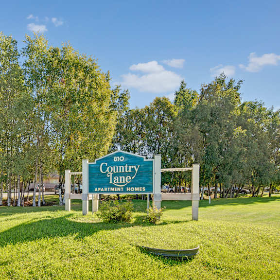 sign reading "810 country lane apartment homes" stands on a lush, green lawn with a backdrop of tall trees under a clear blue sky, conveying tranquility.