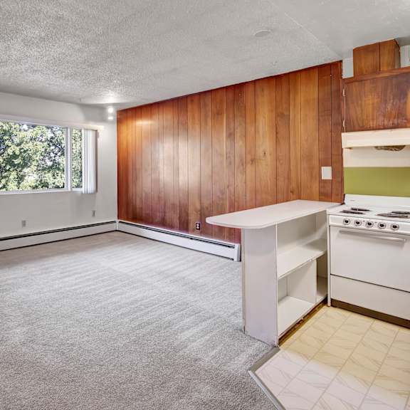 empty living room here at country lane apartments with gray carpet, wood-paneled accent wall, and a large window showing greenery. a compact kitchen with a stove is visible to the right.